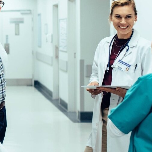 Ein Arzt und eine Ärztin unterhalten sich freundlich mit einer Krankenschwester, im Flur eines Krankenhauses